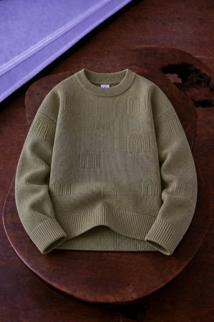 Flat lay of olive green knit sweatshirt with repeated embossed monogram patterns on wooden surface.