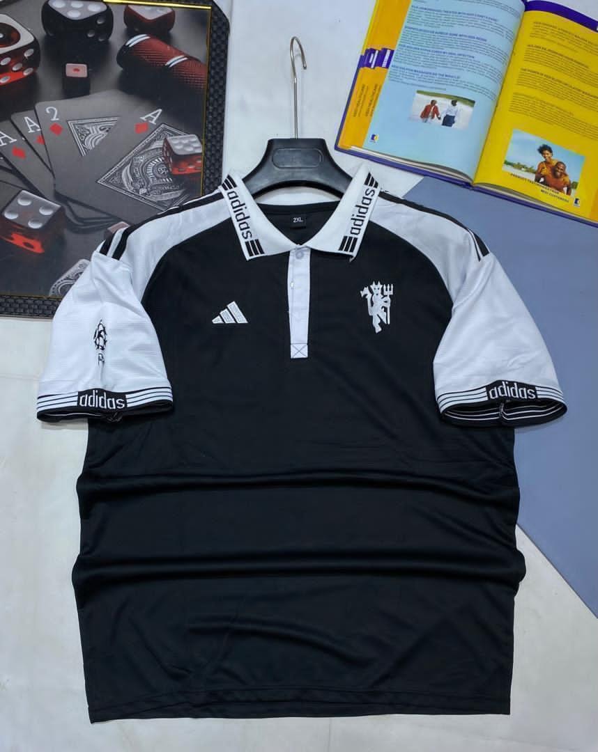 Black and white Manchester United Adidas polo football jersey displayed on a hanger with accessories in background.