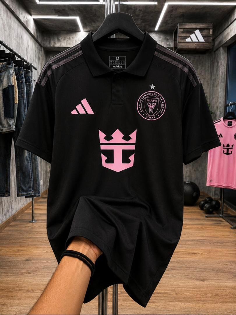 Black Inter Miami polo jersey held by hand showing pink logos and size tag.