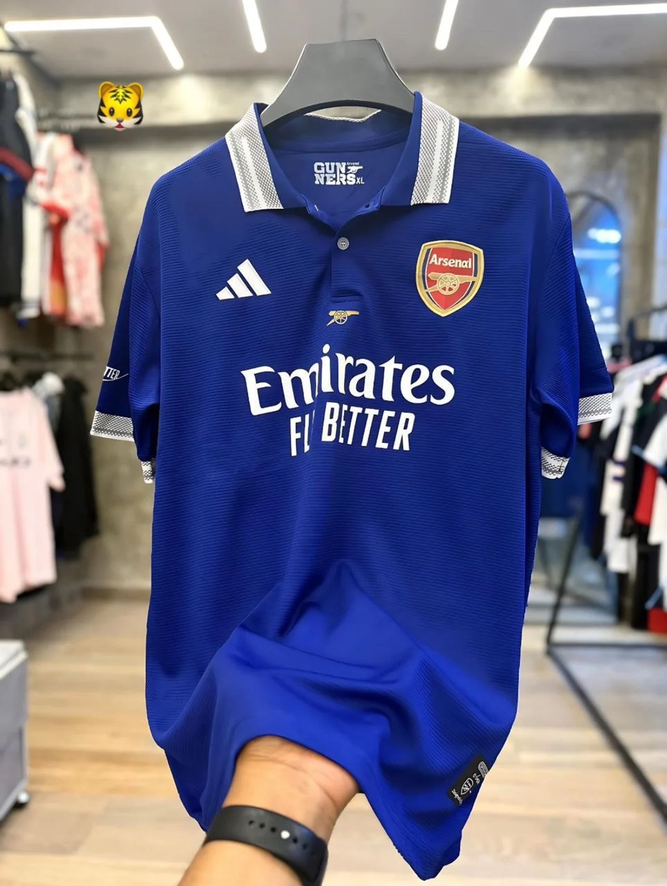 Blue Adidas Arsenal polo jersey with Emirates sponsor on hanger.