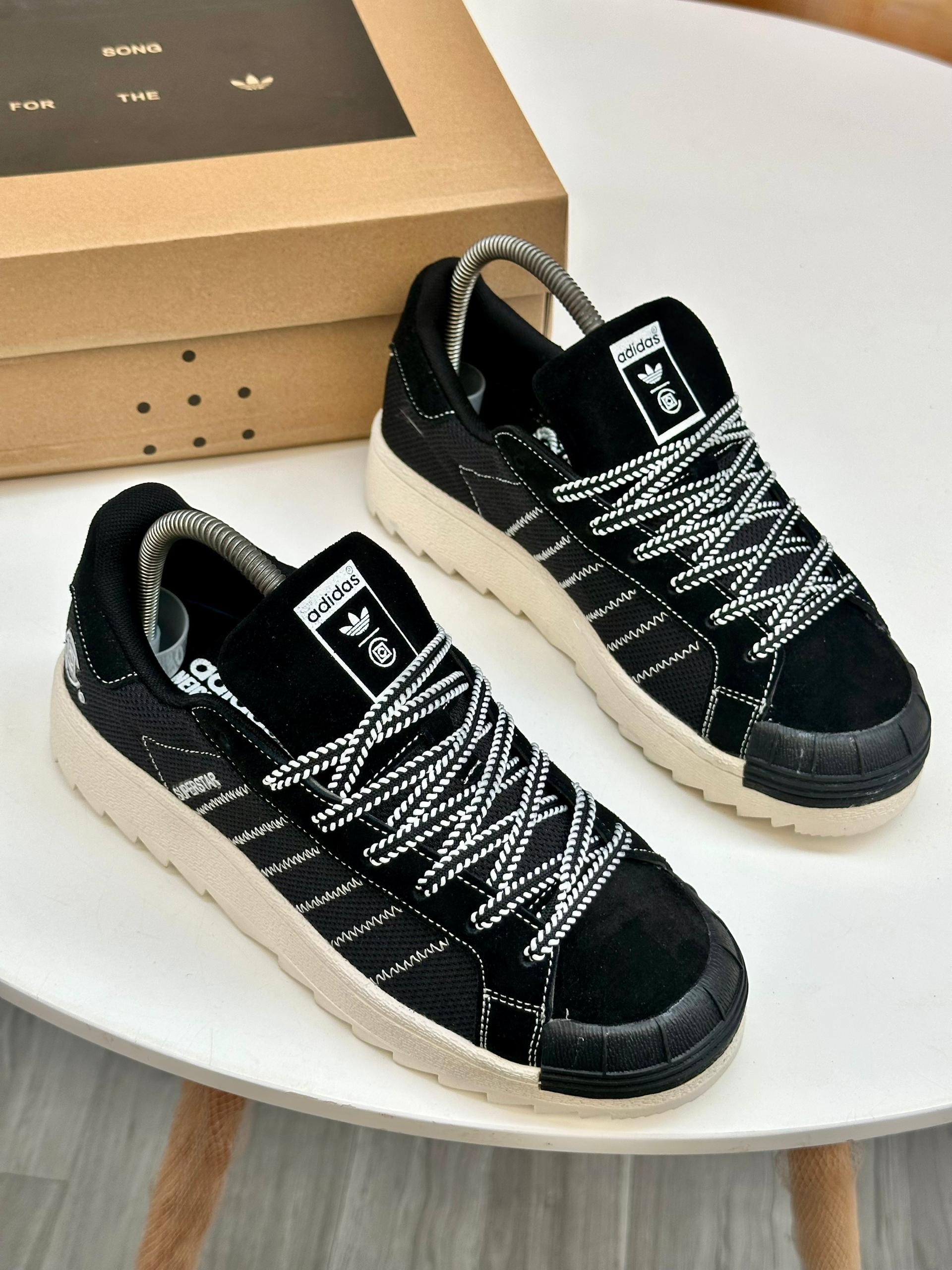 Black Adidas Superstar sneakers with white rope laces and contrast stitching on white table.
