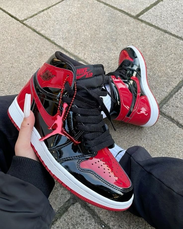 Person holding red and black patent leather Air Jordan 1 High sneakers with red Jumpman charm.
