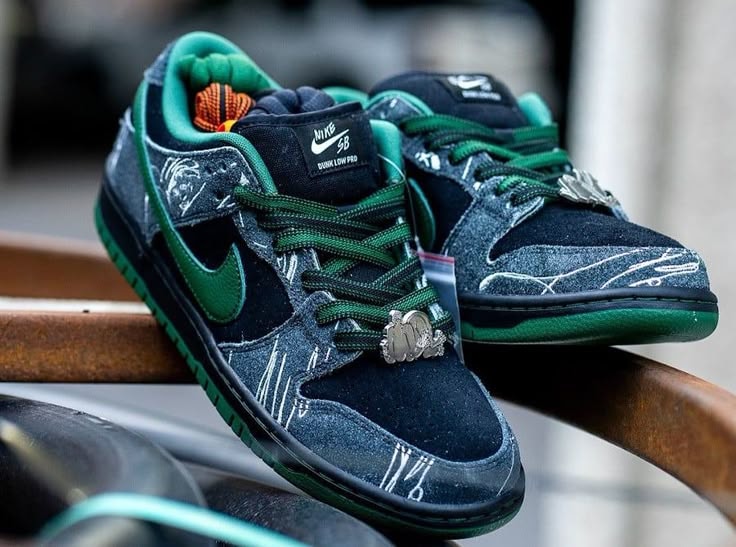 Nike SB Dunk Low in dark blue denim and green with white graphics and metal pendants on wooden rail.