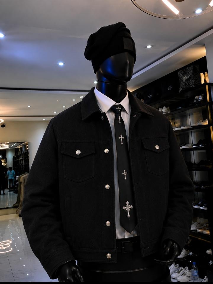 Mannequin wearing black denim trucker jacket with white shirt, cross tie and black beanie in retail store.