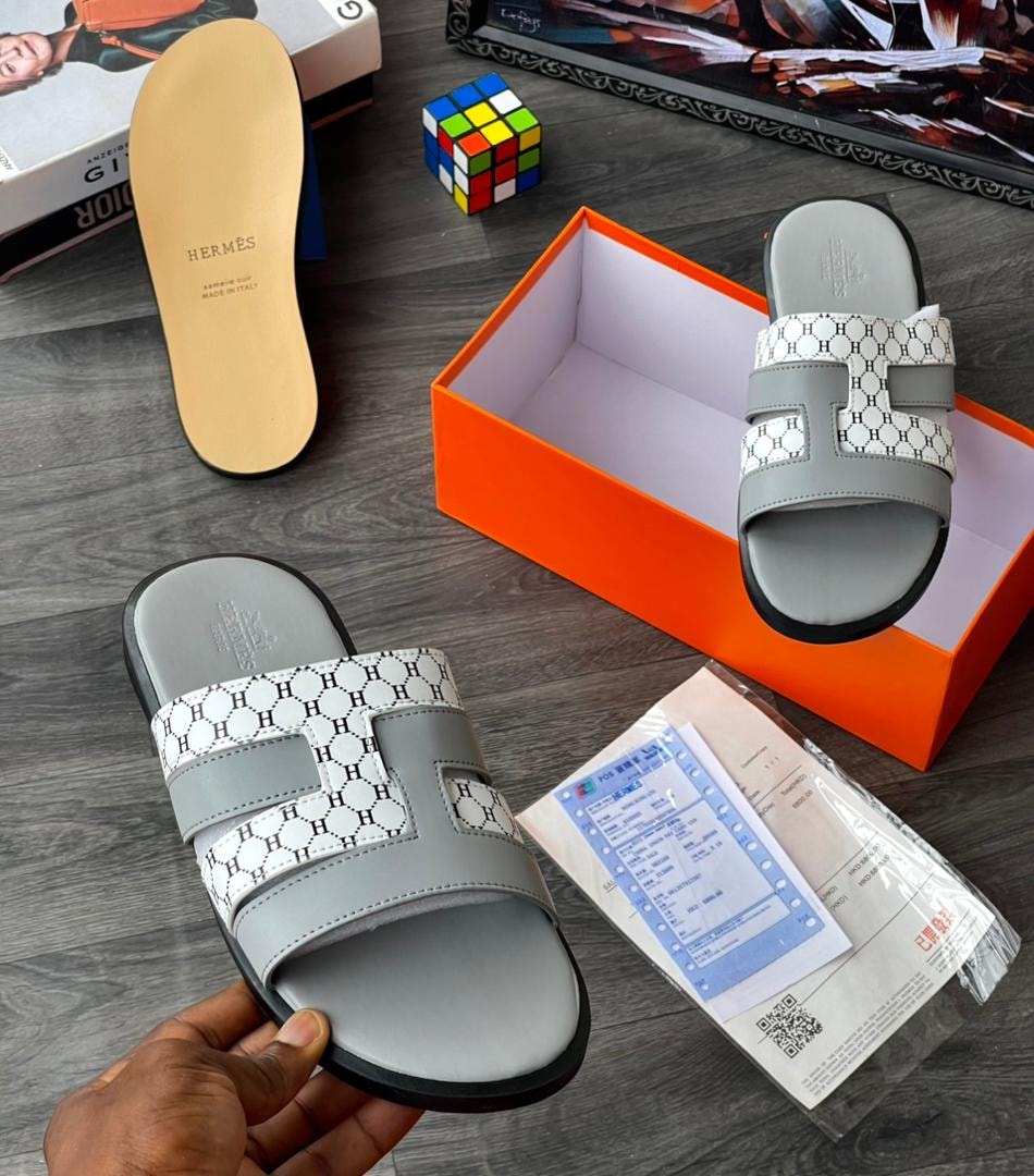 Grey monogram patterned slide sandal with H logos on strap, held in hand with packaging.
