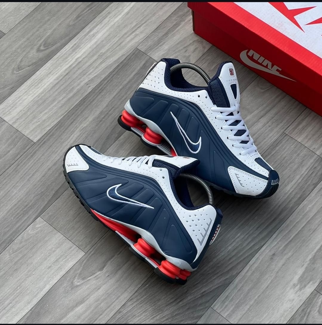 Navy, white and red Nike Shox sneakers on wood floor beside red Nike shoebox.