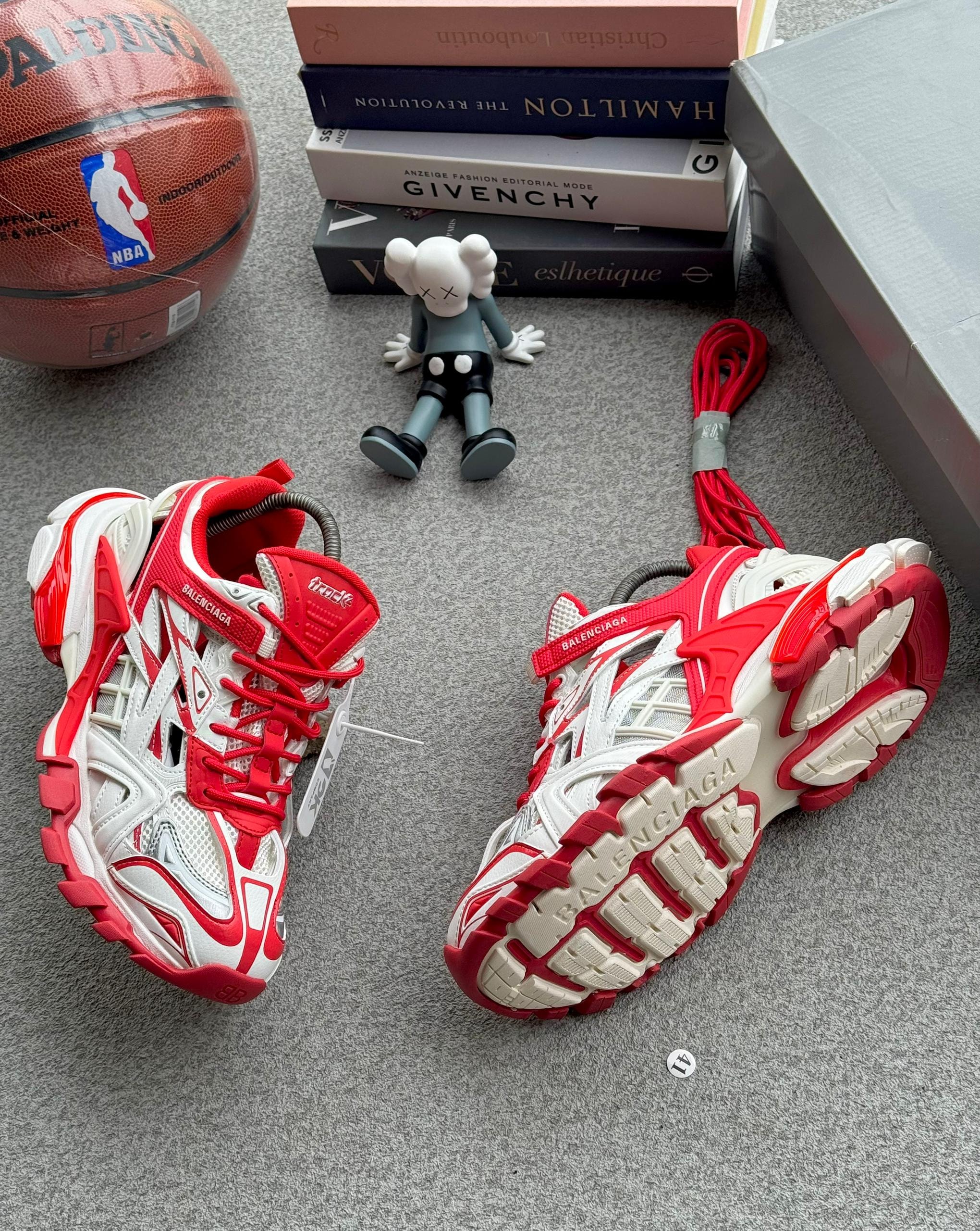 Red and white Balenciaga Track sneakers with accessories, basketball and books on gray surface.