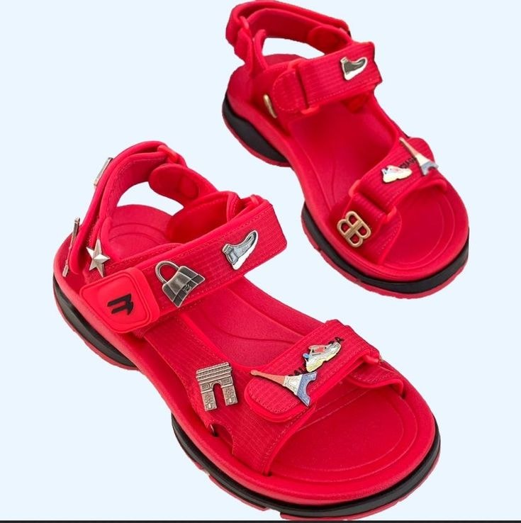 Pair of red sandals featuring multiple decorative metal charms on white background.