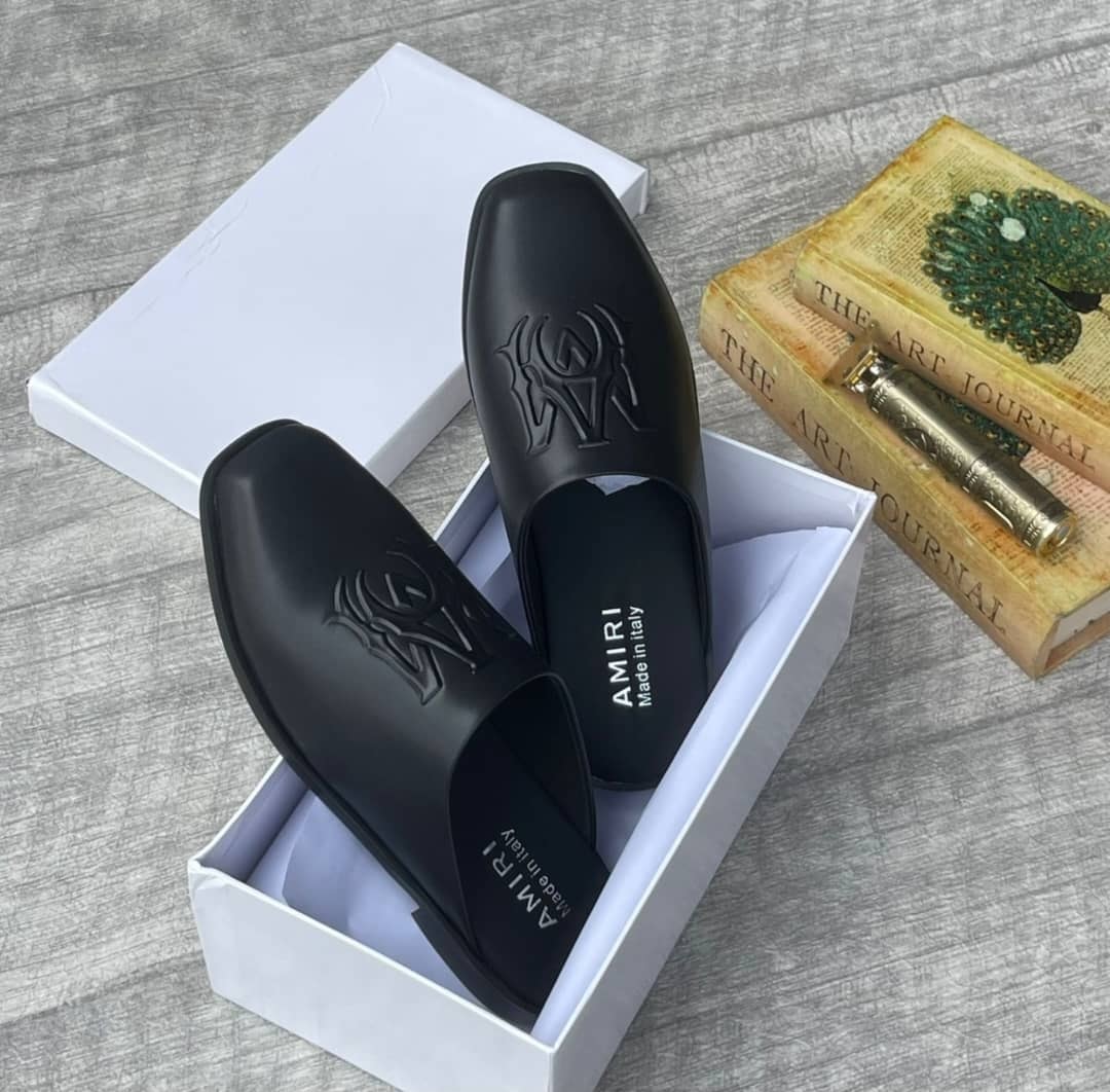 Black Amiri mules with embossed logos in white box surrounded by books and decorative items.