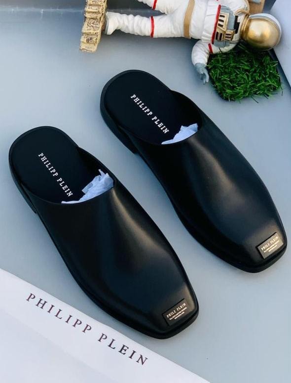Pair of black Philipp Plein mules with branding on insoles and tags, styled with astronaut figure and grass.
