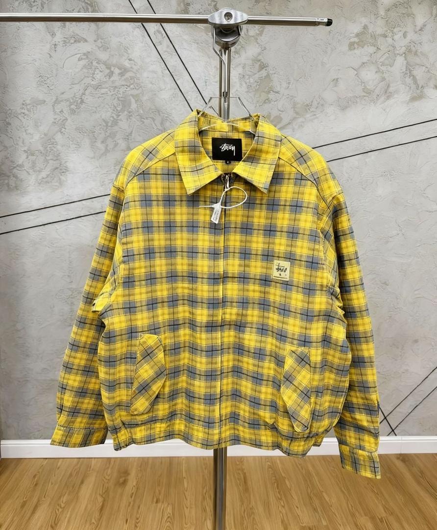 Yellow checkered Stussy jacket hanging on display stand, front view showing yellow and gray plaid pattern.