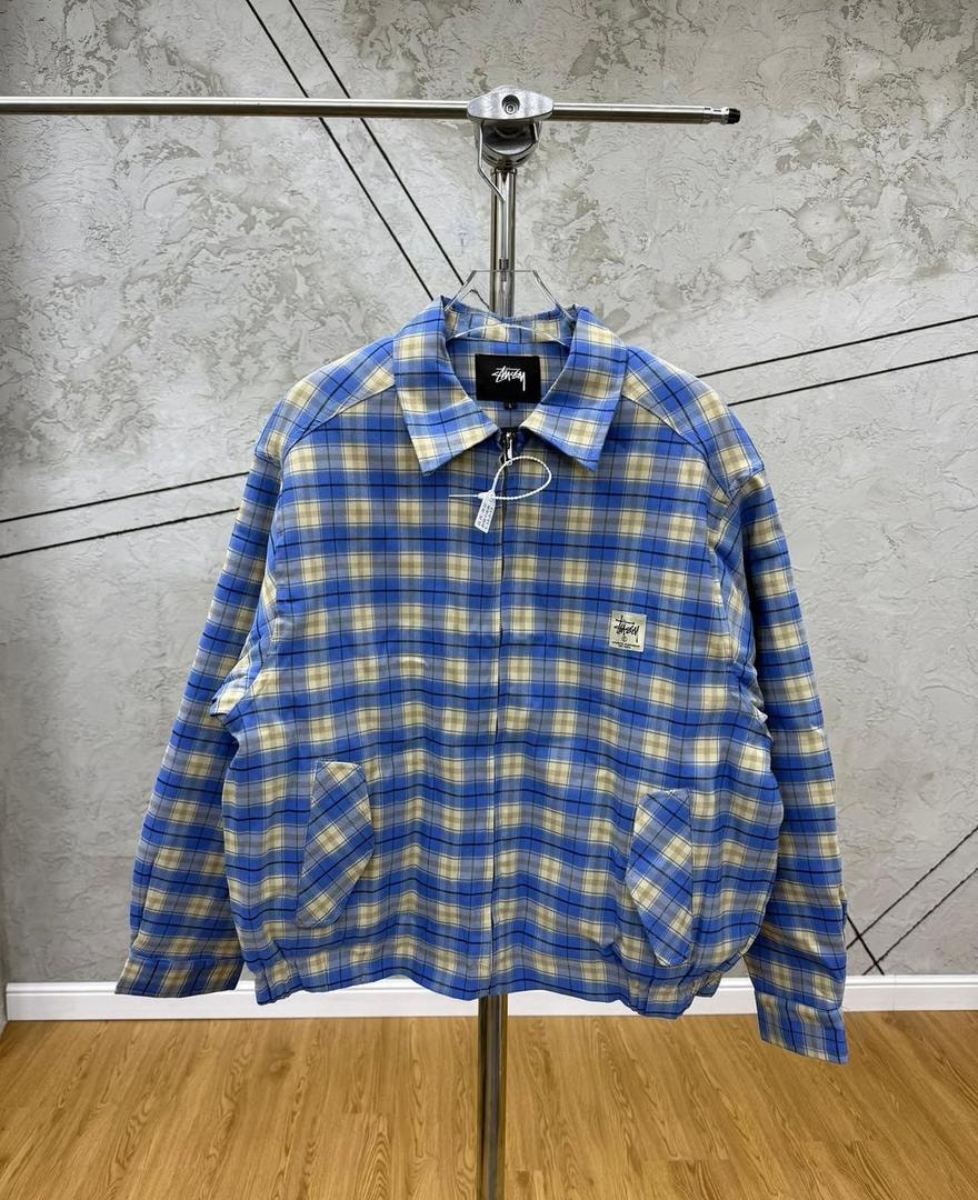 Blue checkered Stussy jacket hanging on display stand, front view showing blue and beige plaid pattern.