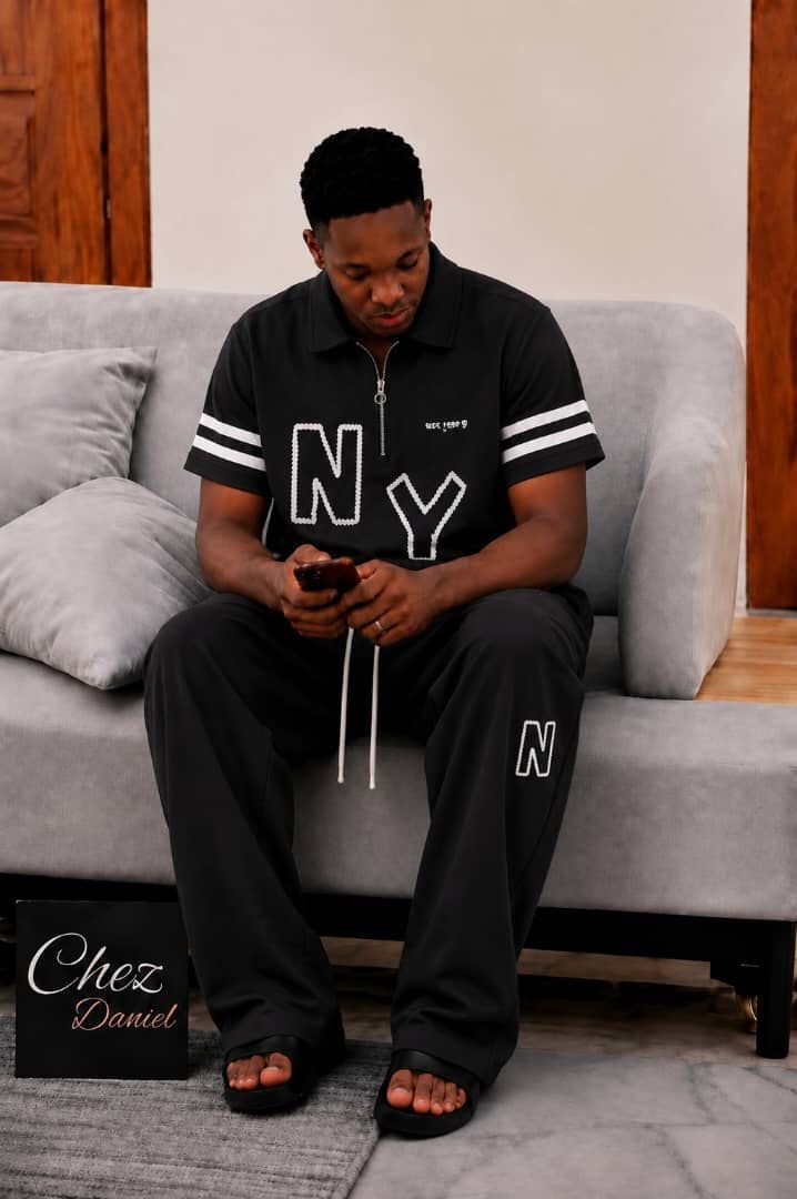Model wearing black NY embroidered 2-piece polo set seated on gray sofa.