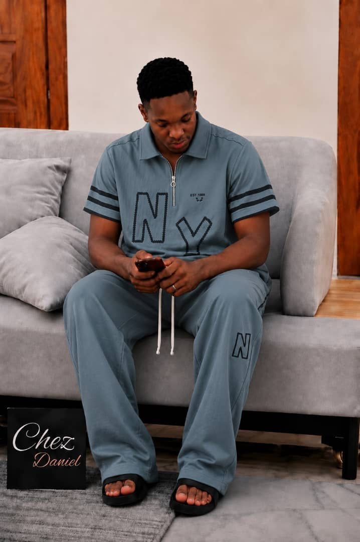 Model wearing light blue NY embroidered 2-piece polo set seated on gray sofa.