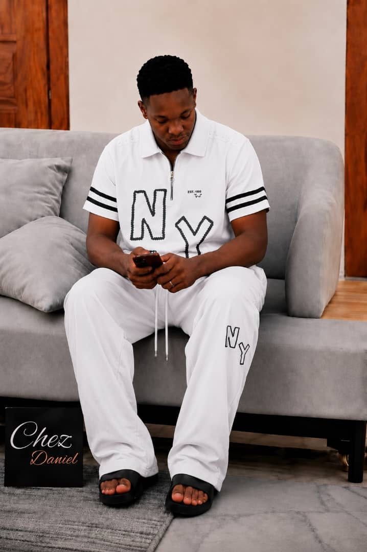 Model wearing white NY embroidered 2-piece polo set seated on gray sofa.