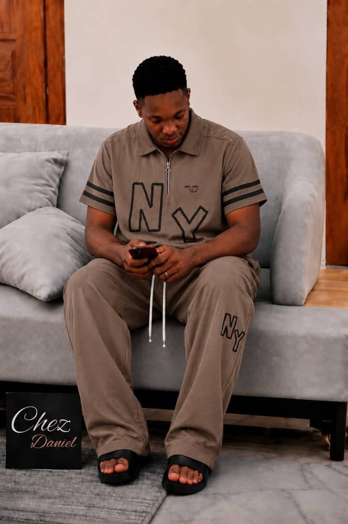 Model wearing brown NY embroidered 2-piece polo set seated on gray sofa.