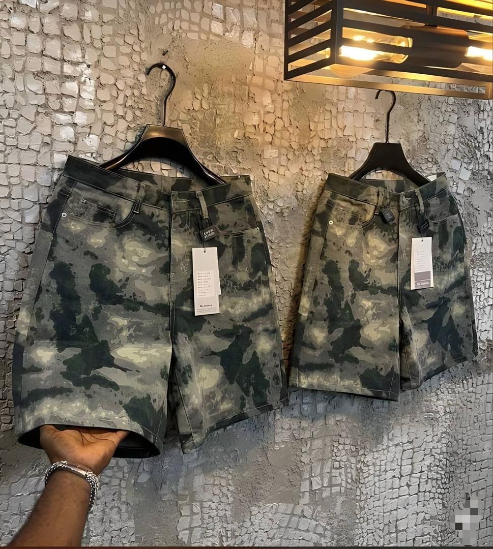 Two pairs of camouflage denim jorts hanging on black hooks against textured wall.
