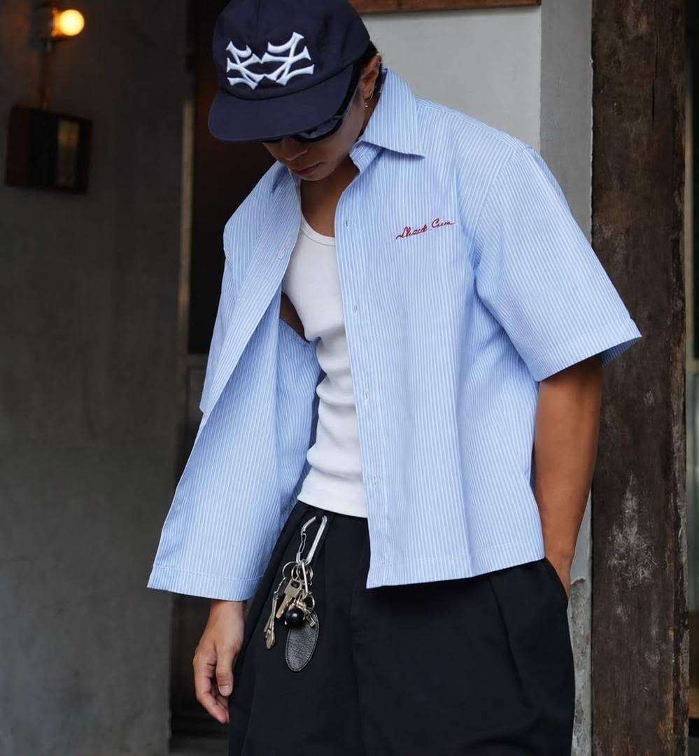 Model wearing light blue pinstriped short sleeve button-up shirt with red chest embroidery.