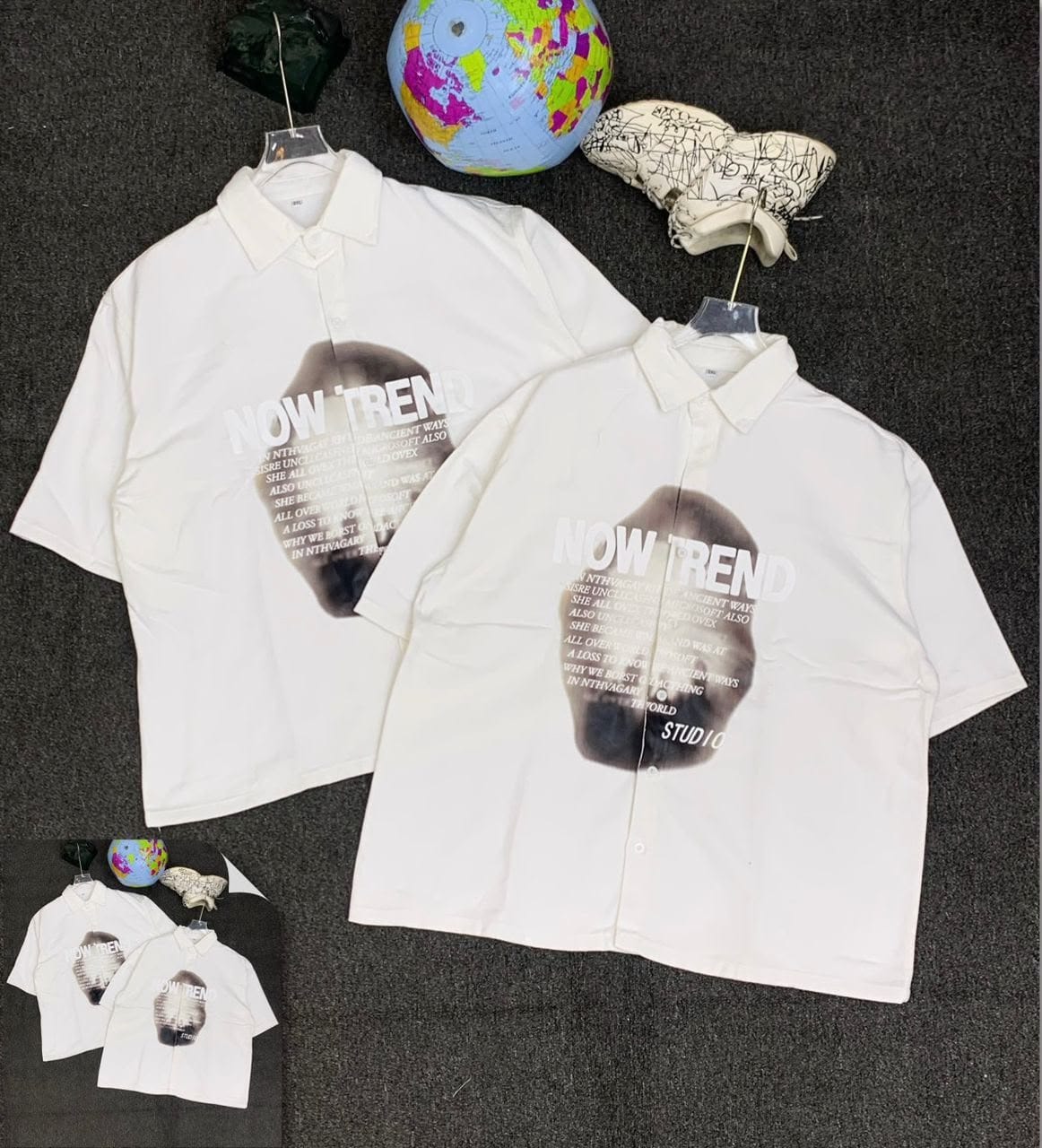 White short sleeve shirts with NOW TREND text print and abstract dark graphic, shown in flat lay with props.