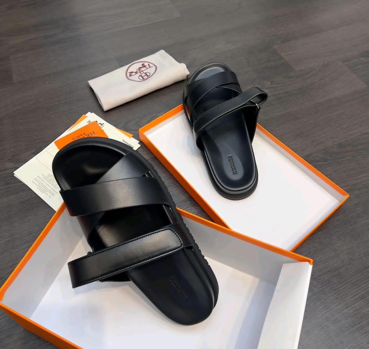 Black Hermes sandals displayed in orange box with white dust bag.