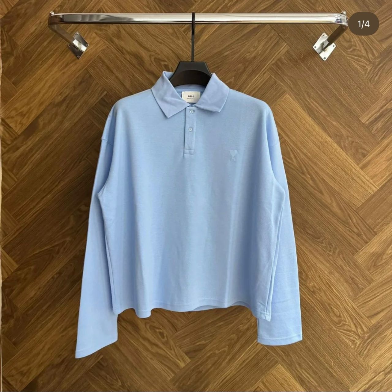 Light blue AMI long sleeve polo shirt hanging on rack against wood herringbone floor.