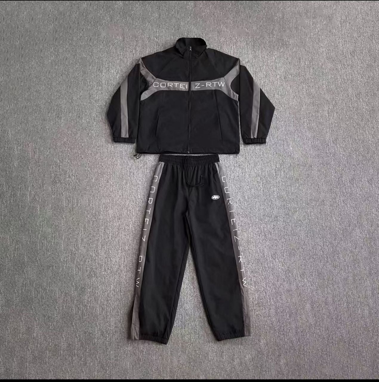 Black and gray Corteiz Z-RTW tracksuit set with zip jacket and jogger pants laid flat.