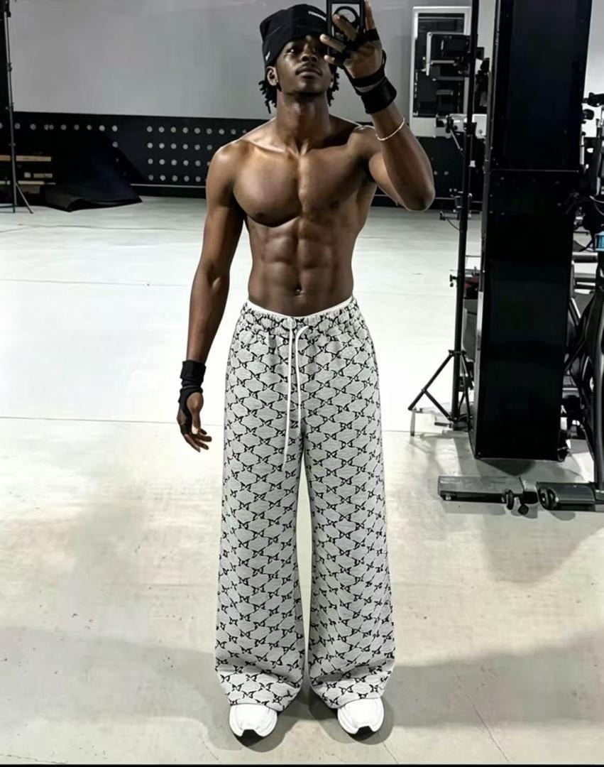 Shirtless model in white monogram print joggers and black cap posing in studio.