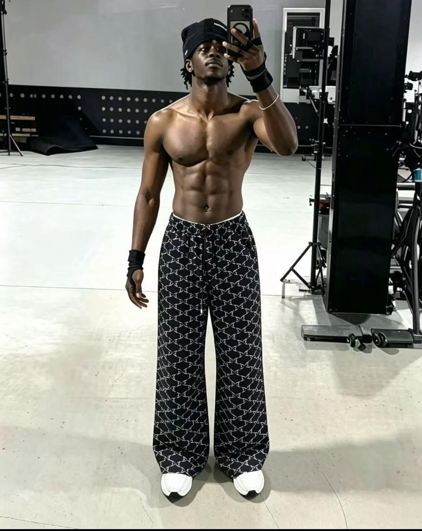 Shirtless model in black monogram print joggers and black cap posing in studio.