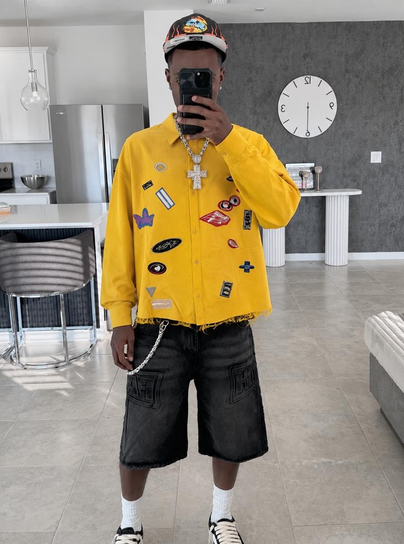 Model in yellow patch shirt with black shorts and chain necklace indoors.