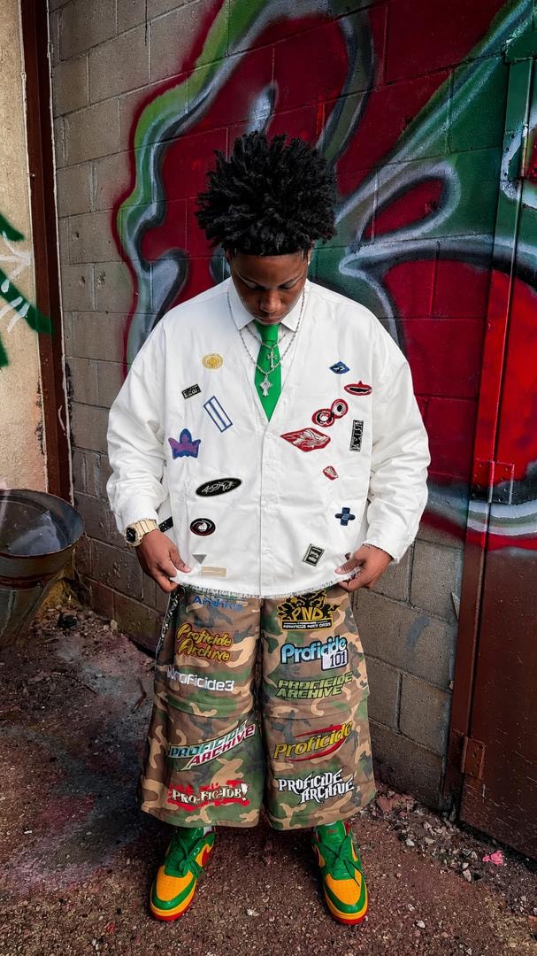 Model in white patch shirt with green tie and camo pants against graffiti wall.
