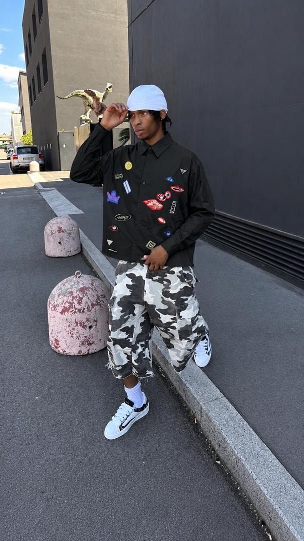 Model wearing black patch shirt with camo shorts and white cap outdoors.