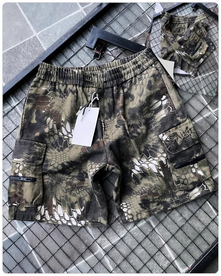 Flat lay of olive reptile print cargo shorts on tiled floor with tag and accessories.