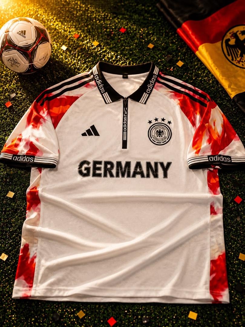 White Adidas Germany football jersey with red patterns, eagle crest and GERMANY text on grass with soccer ball.