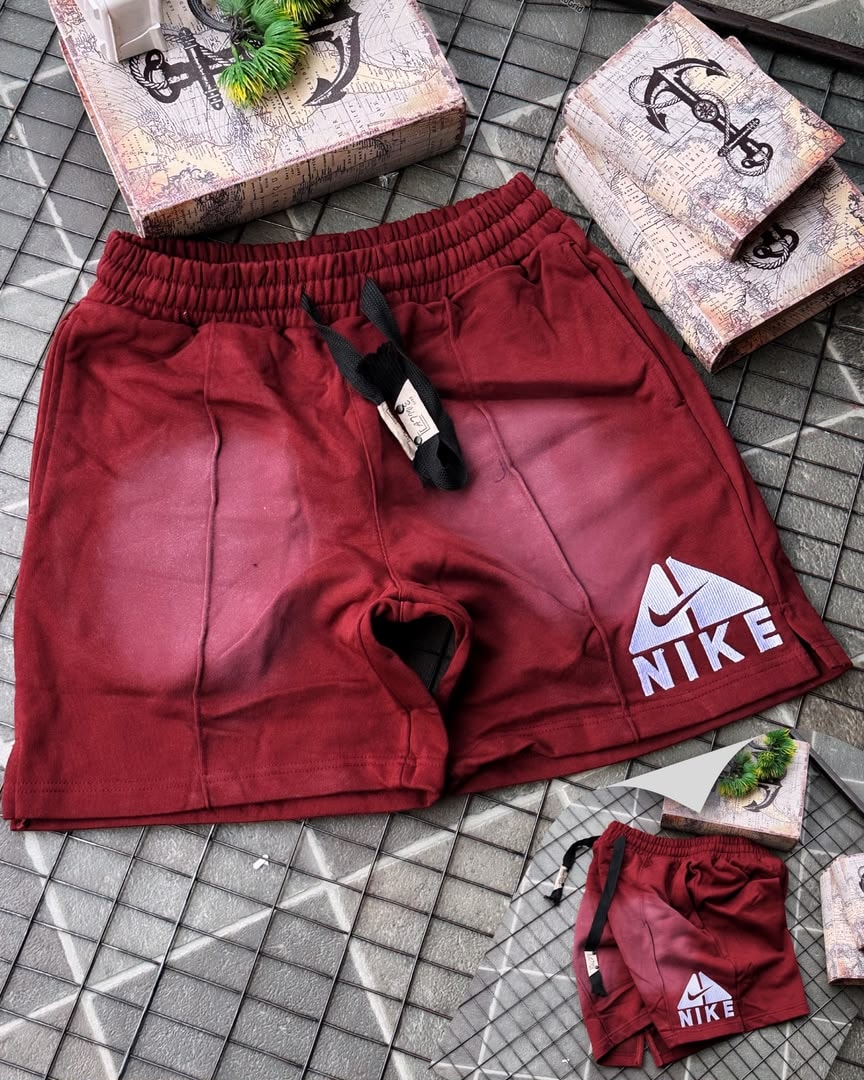 Burgundy Nike fleece shorts with faded gradient and embroidered logo, shown with miniature pair on tiled surface.