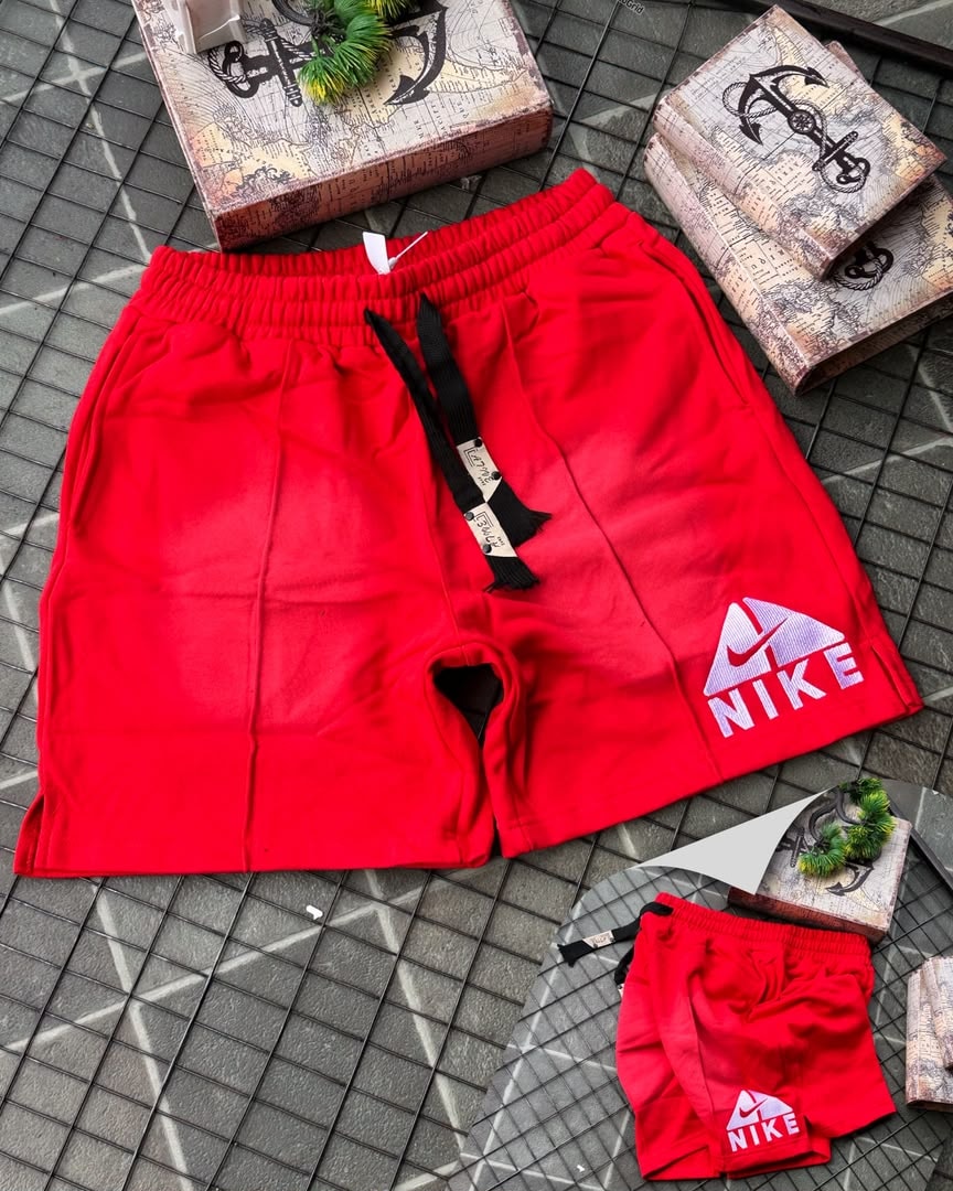 Red Nike fleece shorts with faded gradient and embroidered logo, shown with miniature pair on tiled surface.