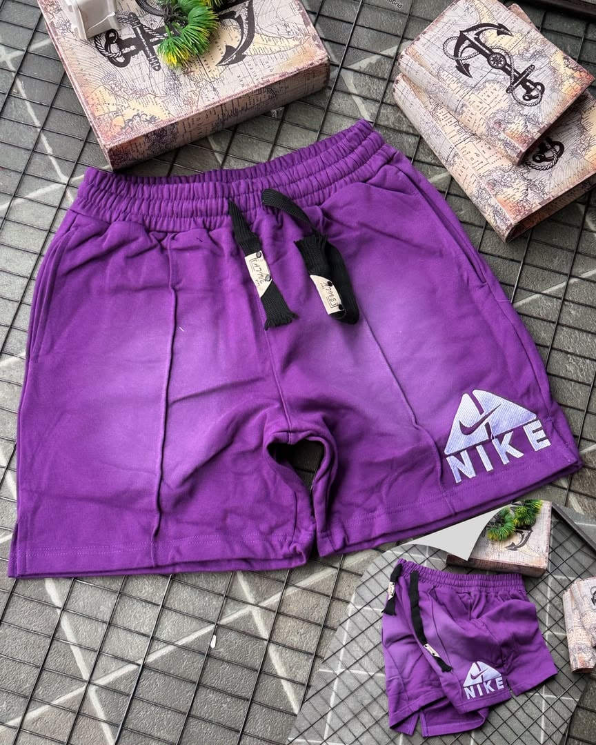 Purple Nike fleece shorts with faded gradient and embroidered logo, shown with miniature pair on tiled surface.