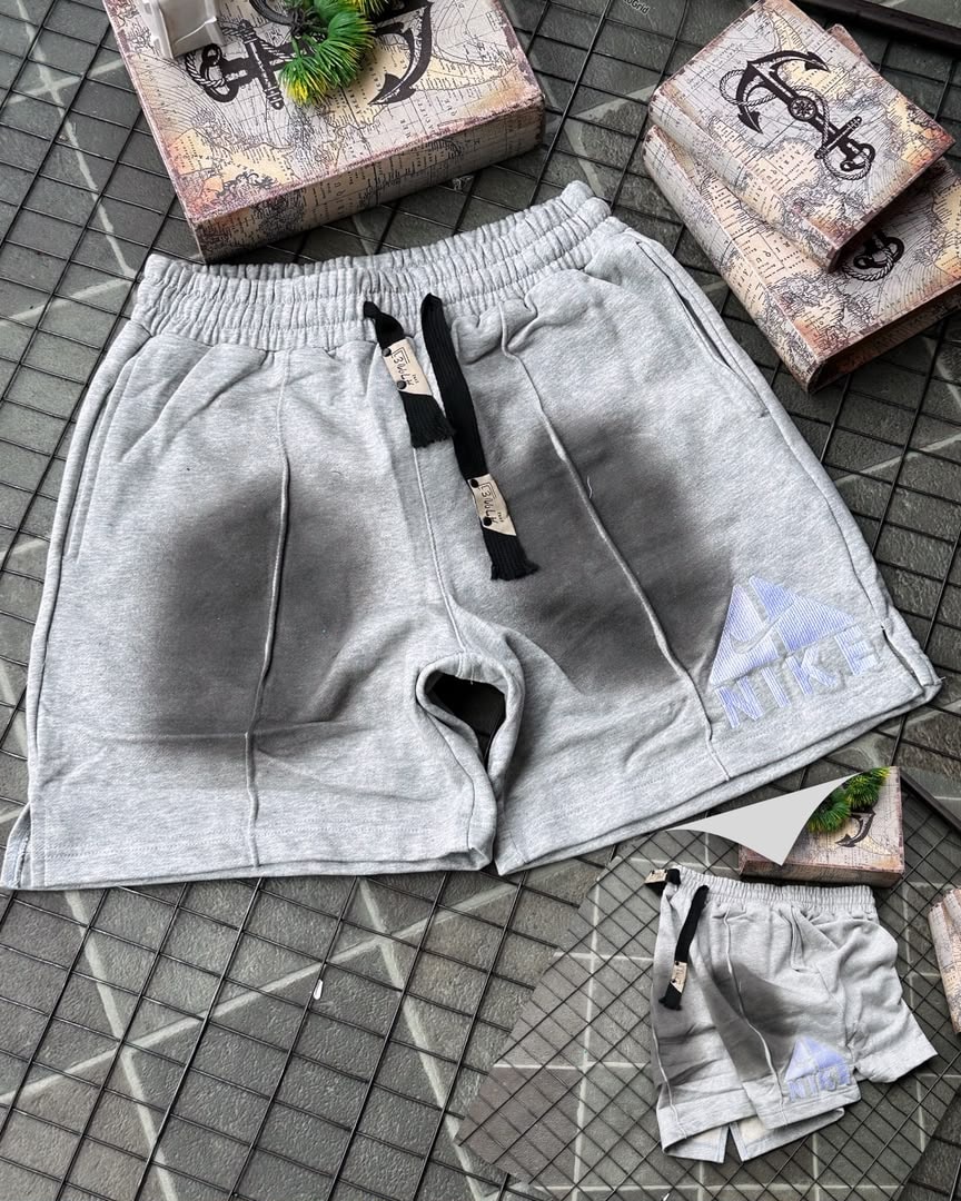 Gray Nike fleece shorts with faded gradient and embroidered logo, shown with miniature pair on tiled surface.