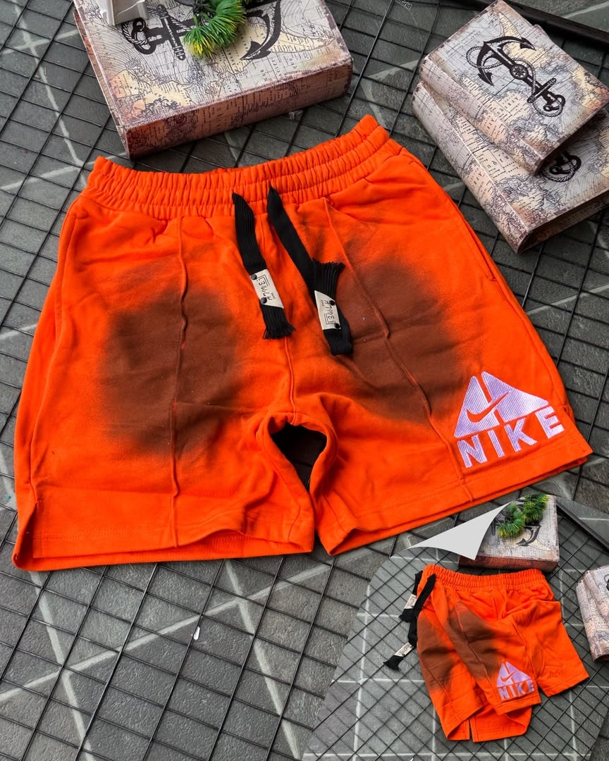 Orange Nike fleece shorts with faded gradient and embroidered logo, shown with miniature pair on tiled surface.