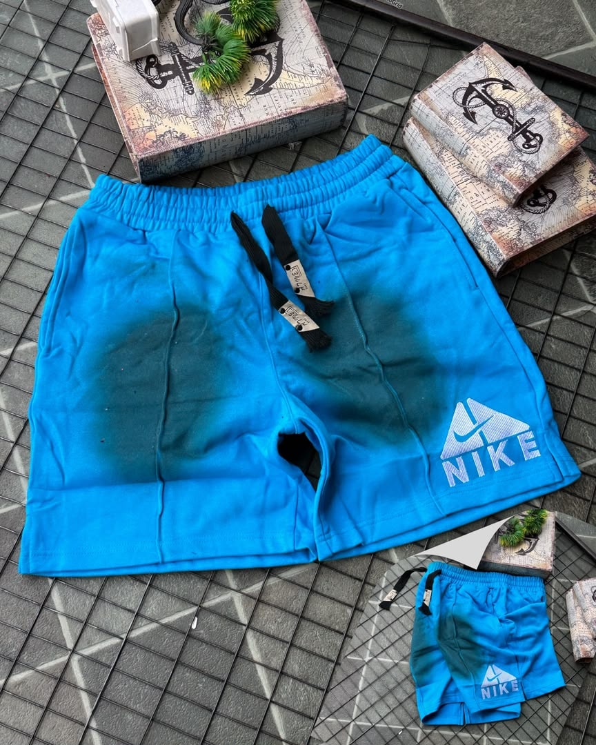 Blue Nike fleece shorts with faded gradient and embroidered logo, shown with miniature pair on tiled surface.