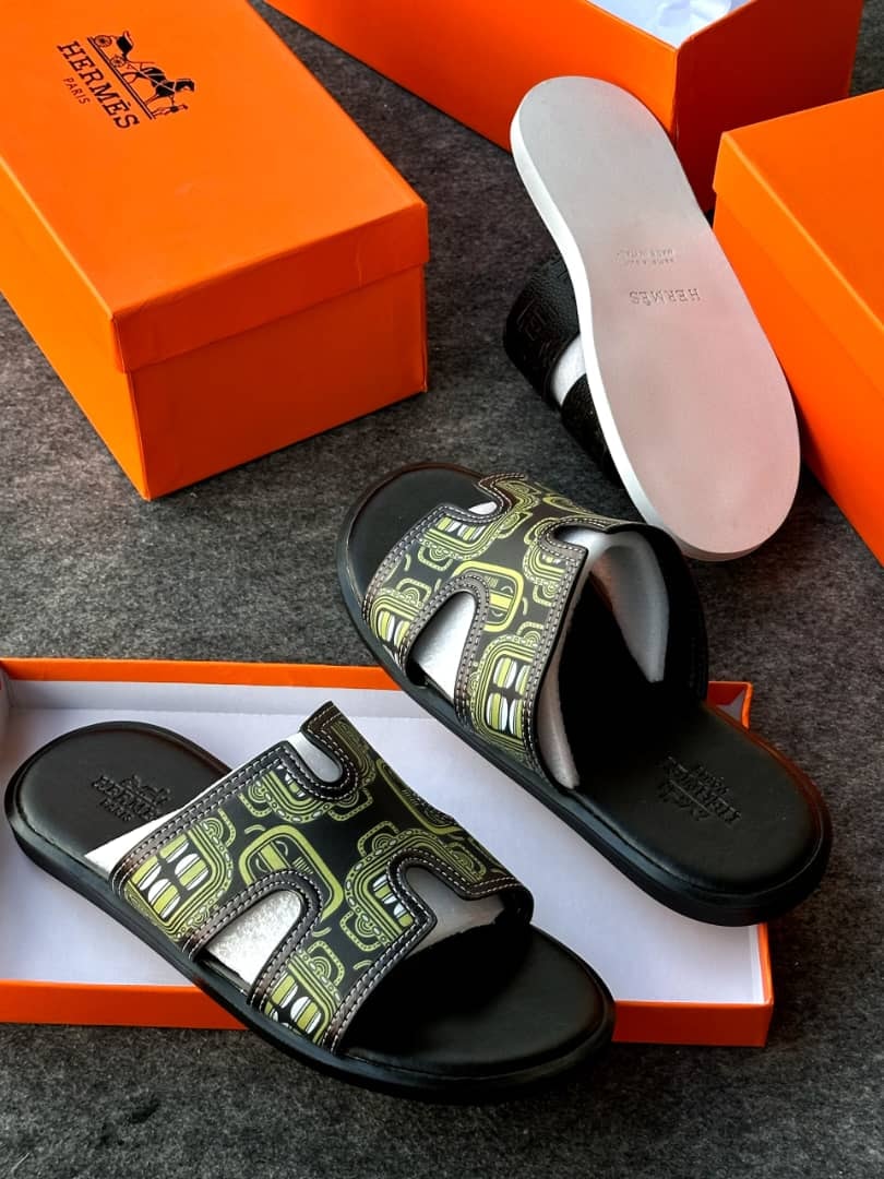 Black slides with lime green geometric pattern and H-shaped strap next to orange Hermes boxes.