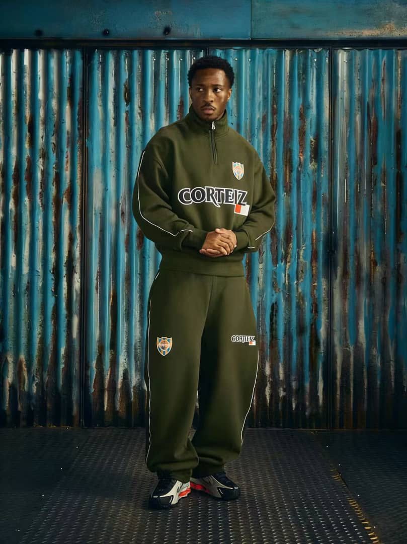 Model wearing olive green Corteiz tracksuit set with logos, posed against blue corrugated metal wall.