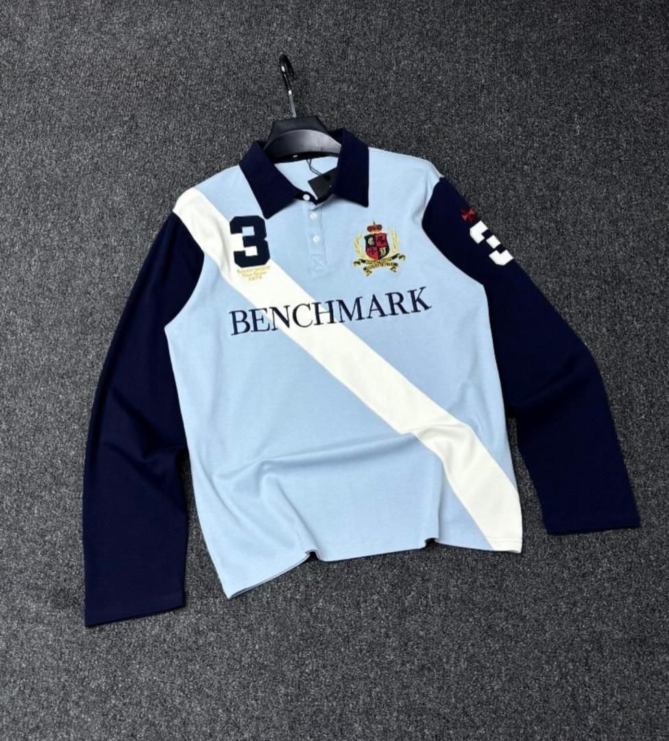 Light blue long sleeve polo shirt with navy contrast sleeves, diagonal stripe, BENCHMARK text and crest logo.