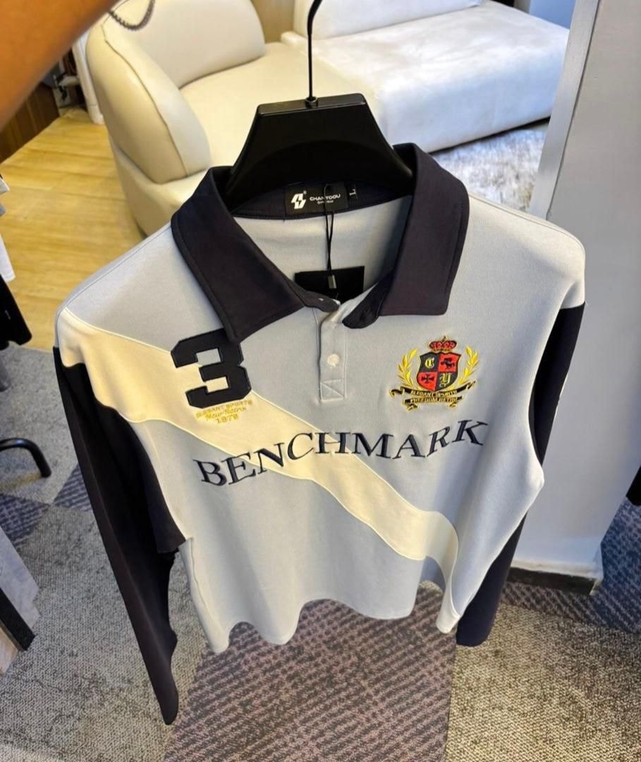 Light gray long sleeve polo shirt with black contrast sleeves, diagonal stripe, BENCHMARK text and crest logo.