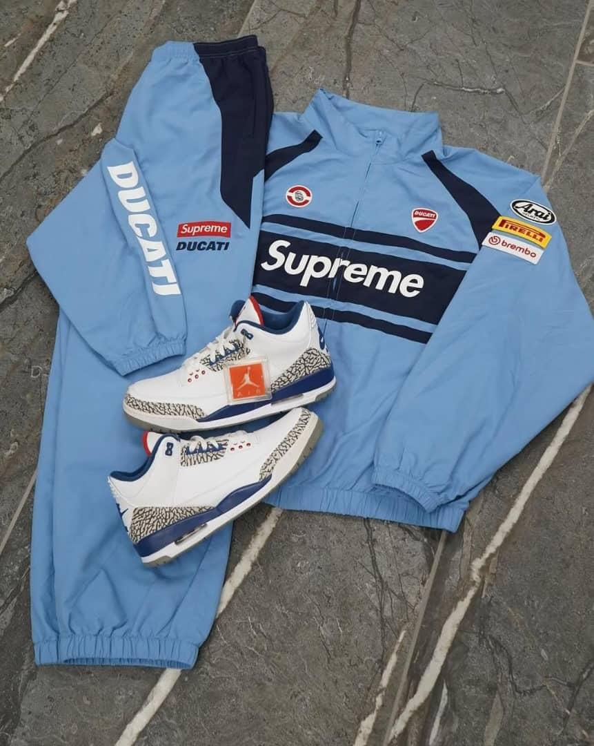 Light blue Supreme Ducati zip-up jacket and track pants with patches, shown with white and blue sneakers.