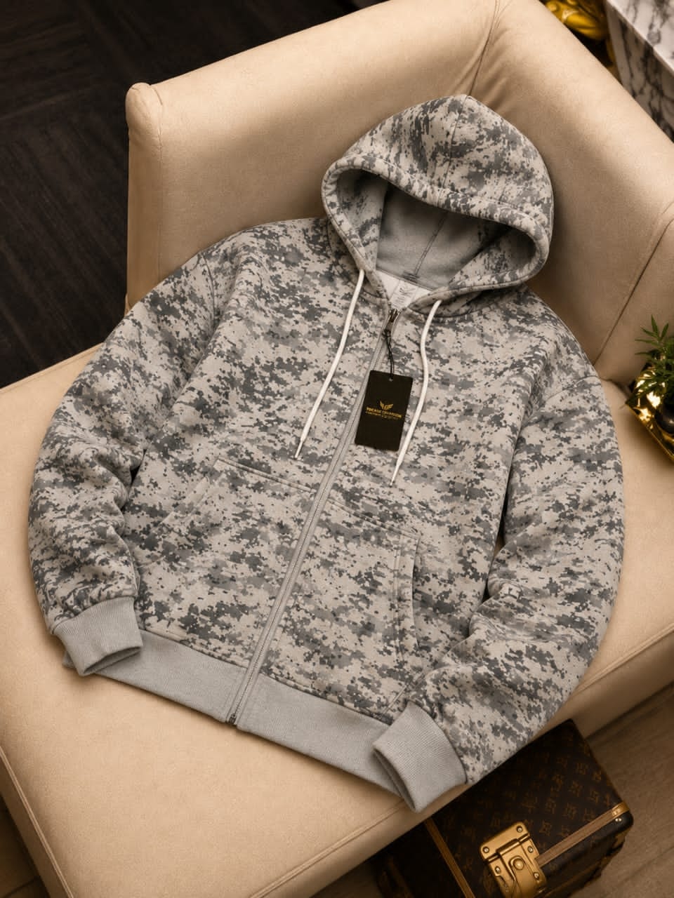Gray digital camouflage zip-up hoodie on a beige chair, showing front view with hood up and black tag.