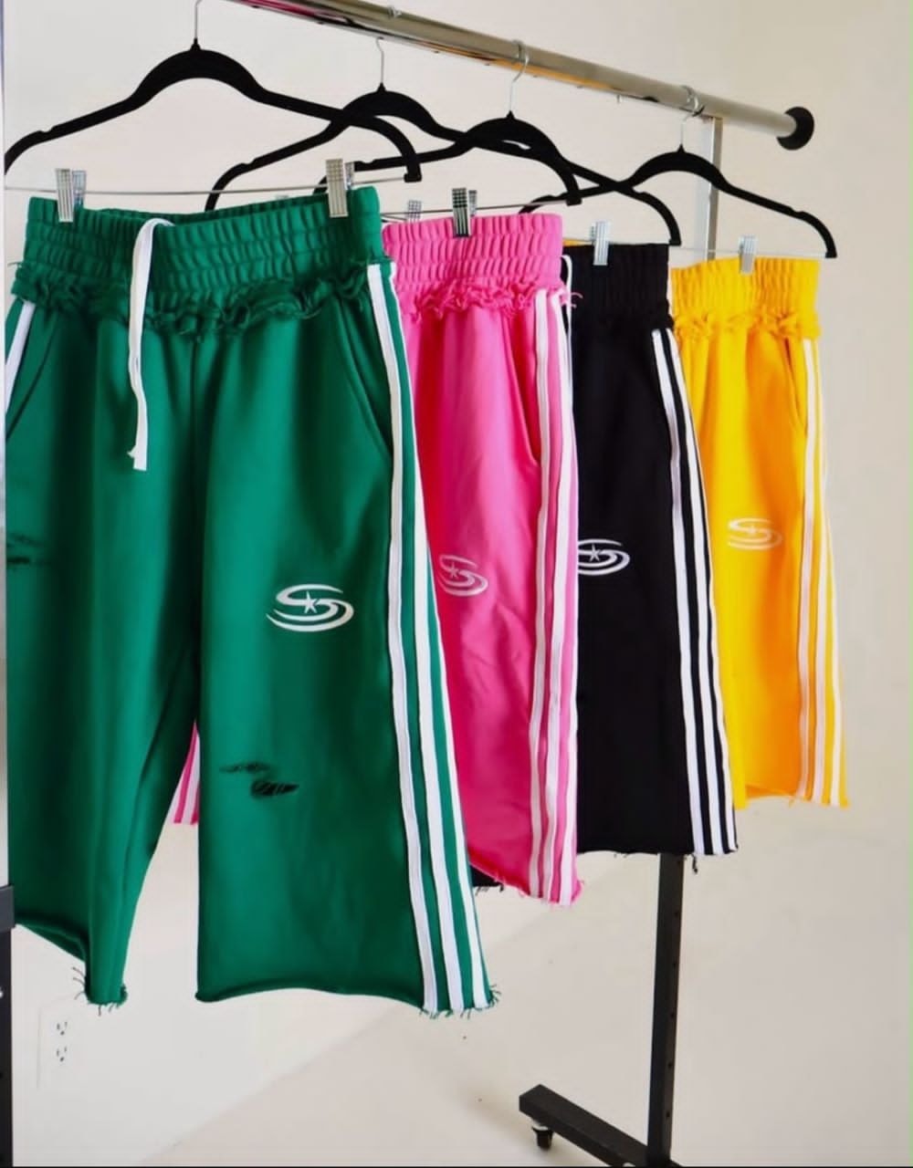 Green, pink, black and yellow striped shorts with embroidered logos hanging on rack.