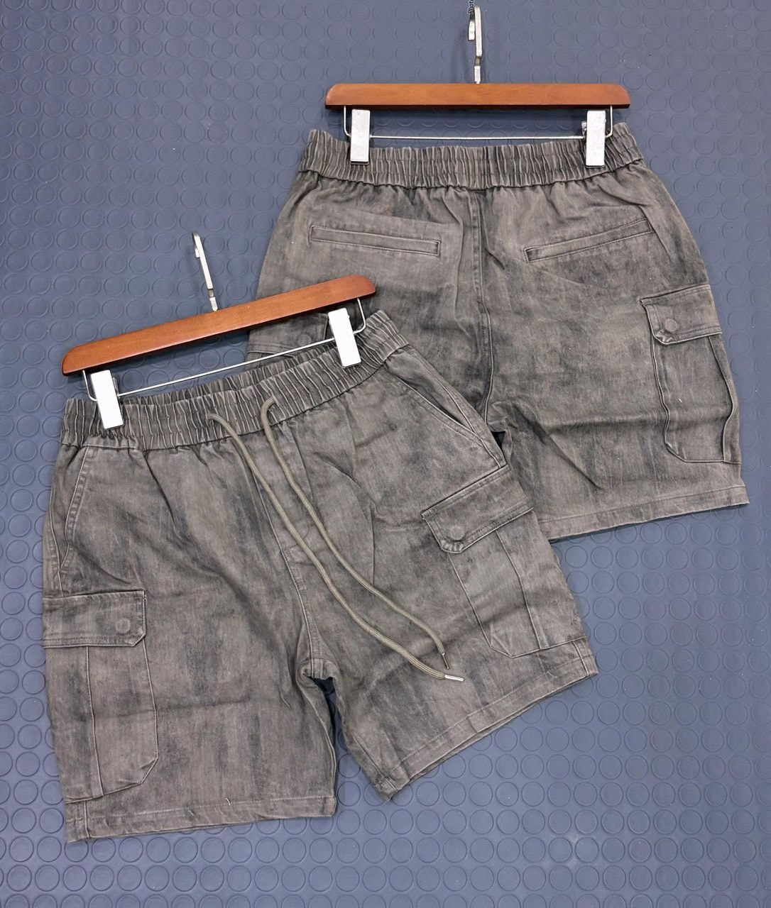 Washed gray cargo shorts hanging on wooden hangers against blue dotted background.