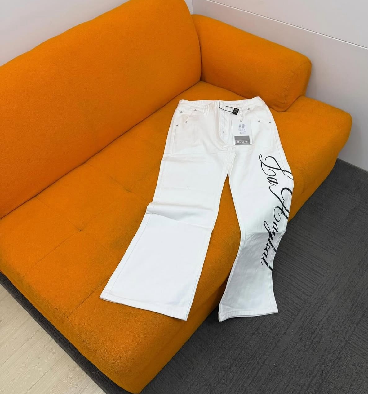 White La Haykal jeans with black script laid flat on an orange couch.
