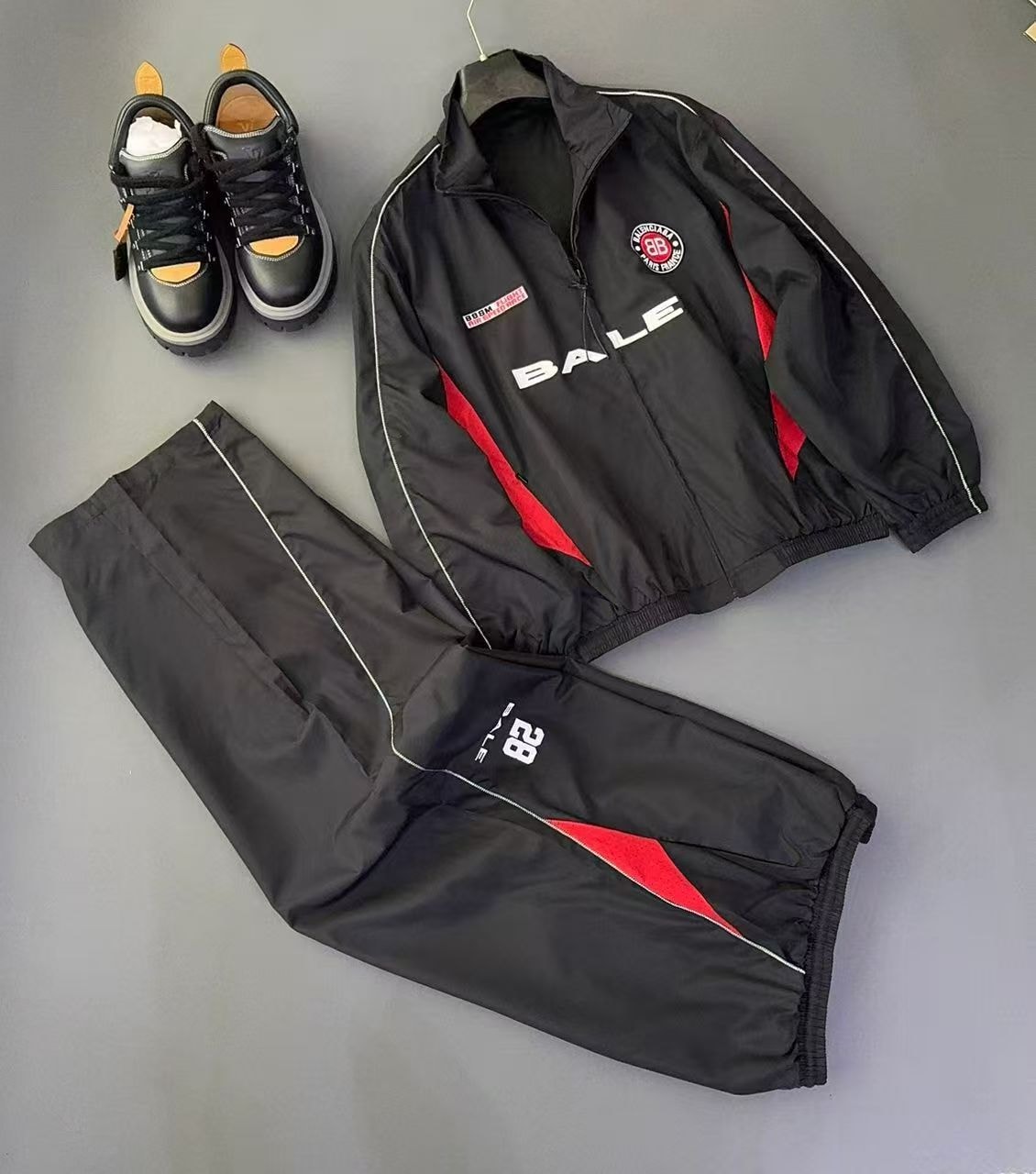 Black tracksuit with red accents, BALE graphics and logos displayed with black and yellow sneakers.