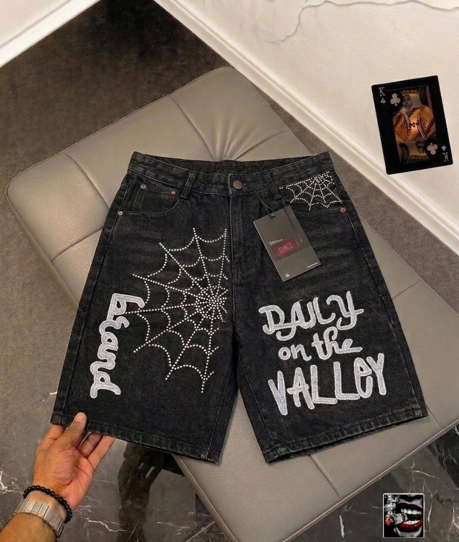 Front view of dark denim jorts with large rhinestone spider web, 'proud' text, 'DAILY on the VALLEY' lettering and back pocket detail.